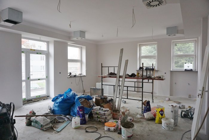 Zdjęcie przedstawia duże remontowane pomieszczenie, w którym stoi małe rusztowanie, sporo farb i narzędzi malarskich